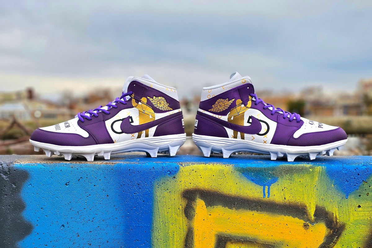 A pair of purple and white high-top football cleats with gold player illustrations, displayed on a graffiti-covered concrete ledge with an outdoor city background.
