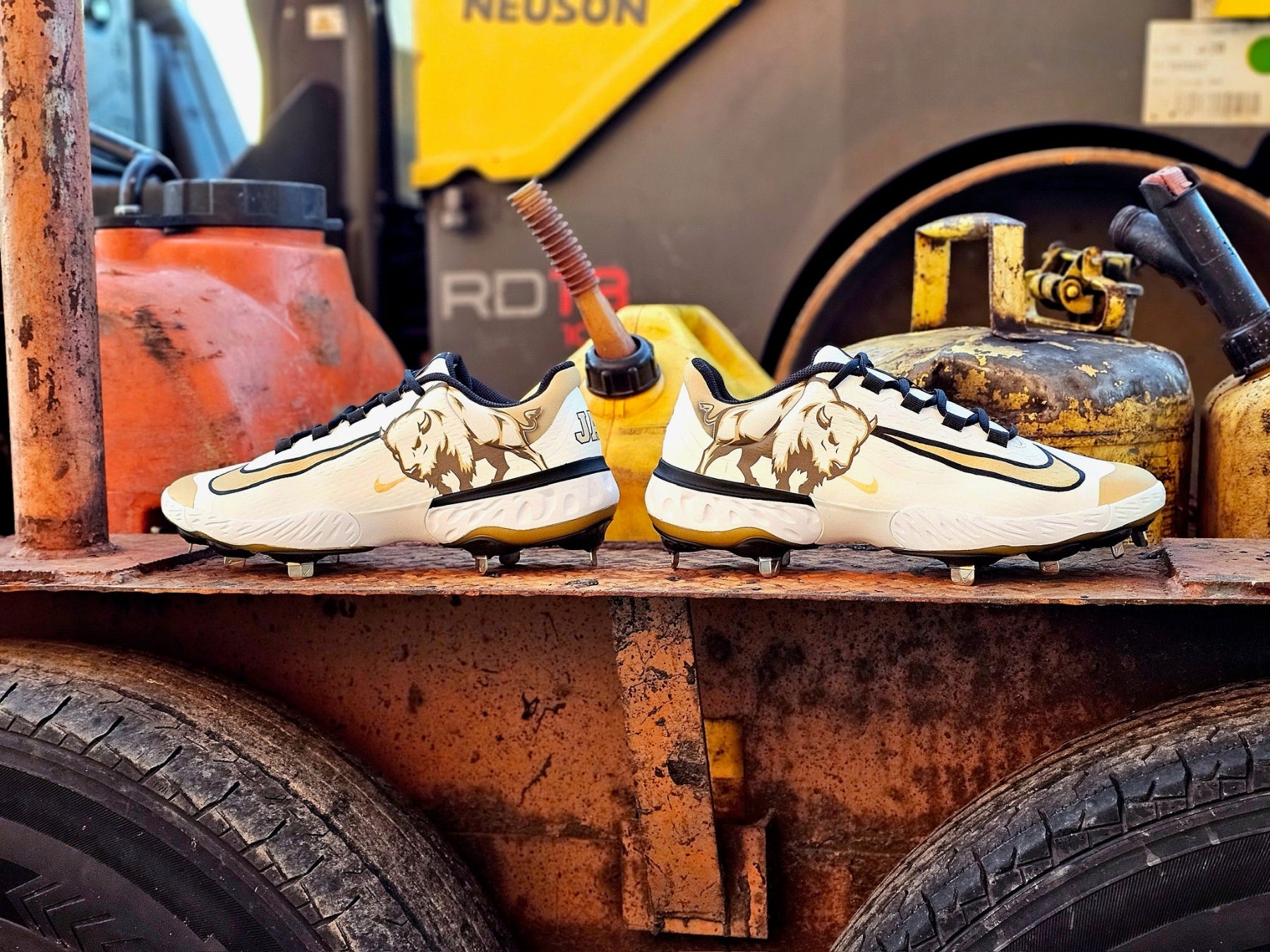 A pair of Nike baseball cleats with a gold and black lion graphic on a rusty metal platform, with heavy machinery and gas cans in the background.