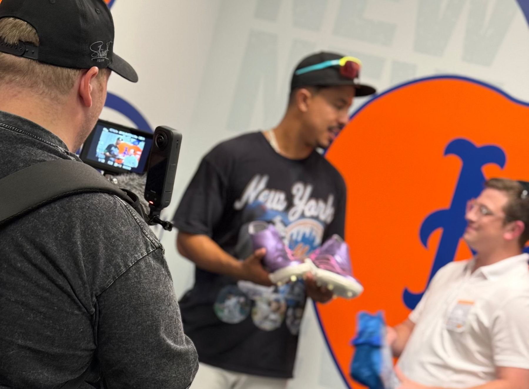 Two men interacting with a third person holding a video camera in front of a Mets logo.