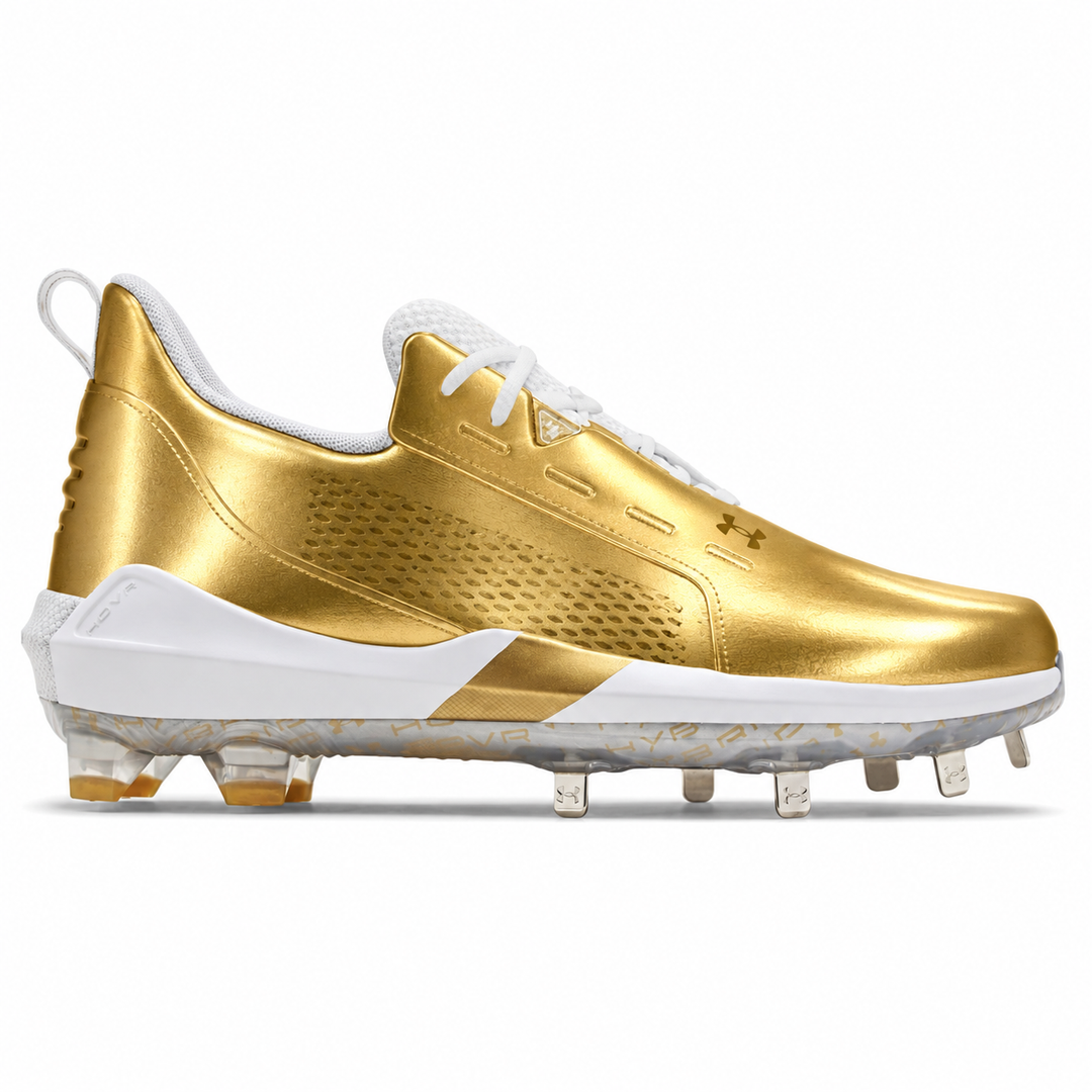A gold and white Under Armour baseball cleat with clear spikes, shown from a side profile on a white background.