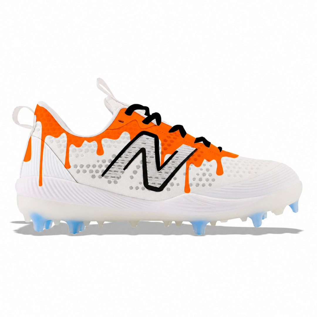 A white New Balance baseball cleat featuring bright orange paint-drip accents, a black outlined 'N' logo, and light blue cleats on a white background.