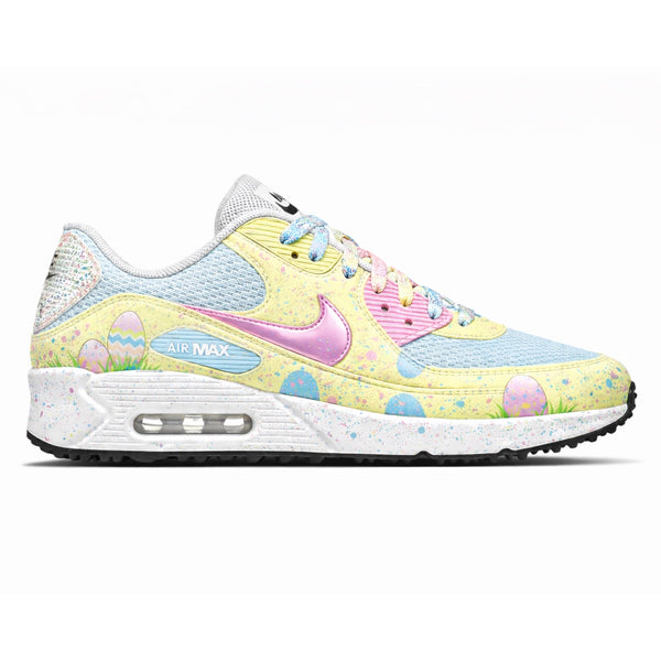 Custom painted Spring Hunt Nike Air Max 90 G golf shoes featuring a pastel yellow base with illustrated Easter eggs and green grass scene along the lower panel, baby blue mesh overlays, a pink glitter Swoosh, pink striped tongue, multicolor speckled midsole, visible Air Max unit, and blue and white laces by Stadium Custom Kicks