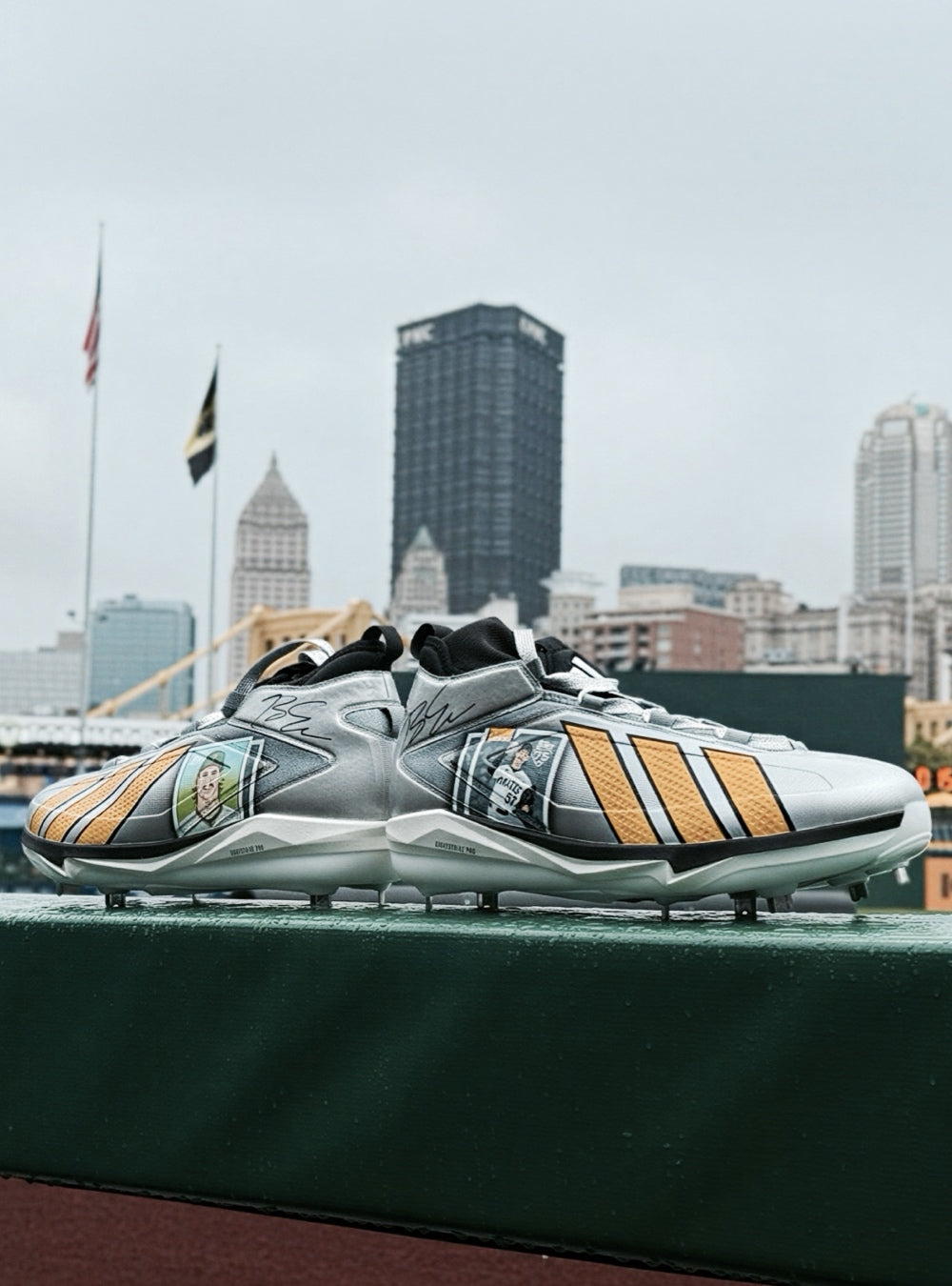A pair of Adidas baseball cleats in metallic silver and golden yellow, displayed on a green field with a city skyline in the background.