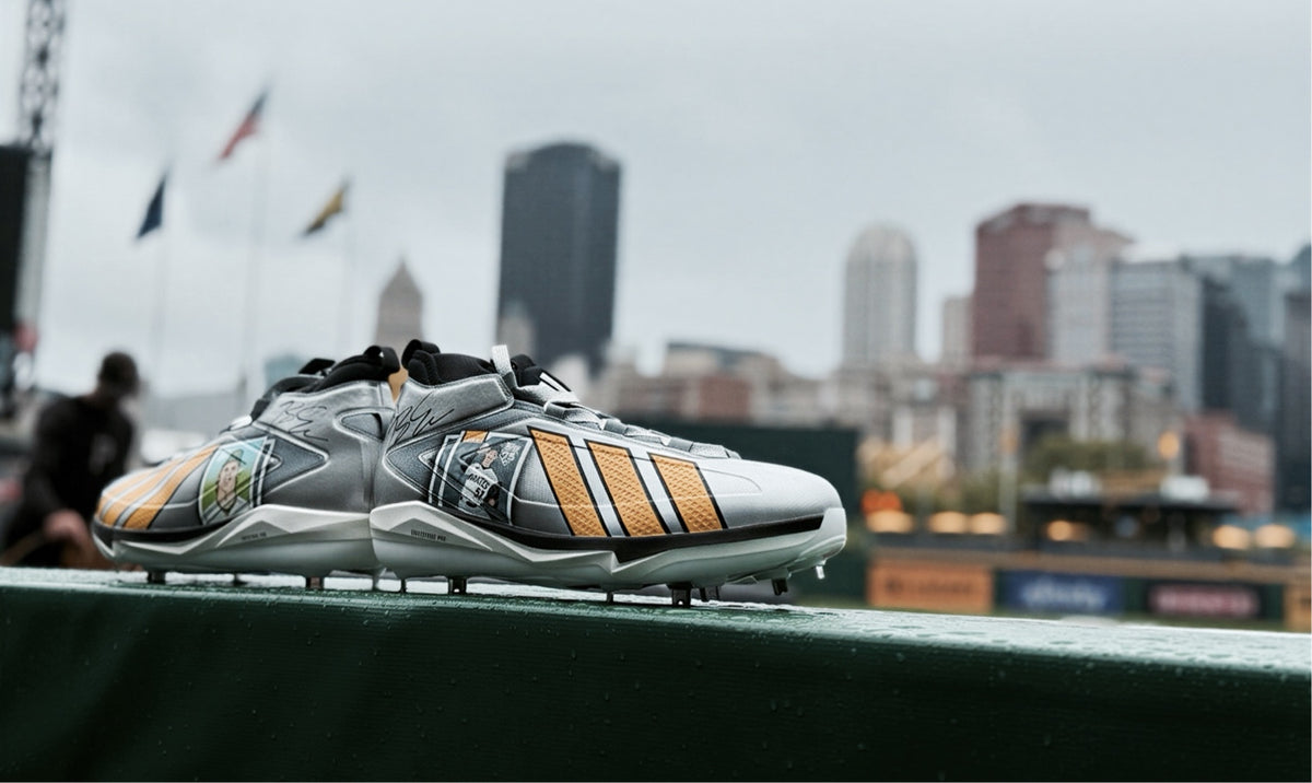 A pair of Adidas baseball cleats in a metallic silver and black colorway with pirates golden yellow stripes, resting on a green field railing with a blurred city skyline in the background.