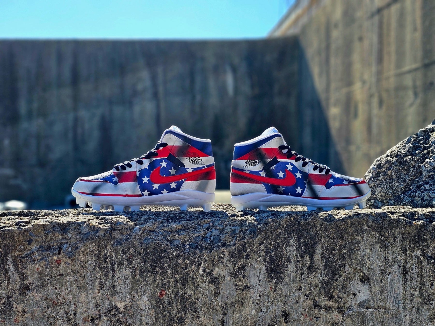 Pair of high-top baseball cleats with American flag design on a concrete ledge.