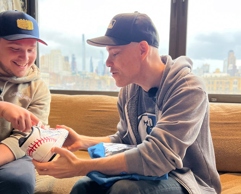 Two men sitting on a couch with a cityscape view, one holding a pair of custom sneakers.
